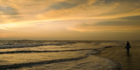 The image captures a serene beach scene with gentle waves lapping against the shore. The sky is filled with warm hues of yellow and orange as the sun sets on the horizon. On the right side, a solitary figure strolls along the wet sand, creating a peaceful and reflective atmosphere.