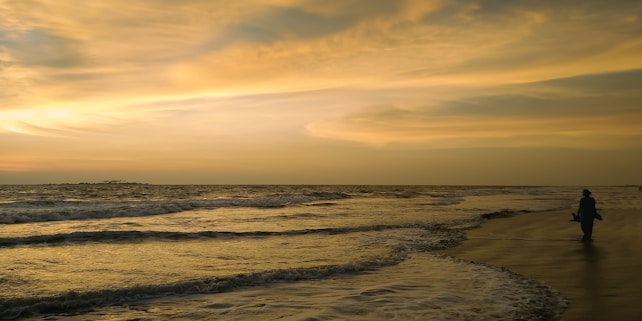 The image captures a serene beach scene with gentle waves lapping against the shore. The sky is filled with warm hues of yellow and orange as the sun sets on the horizon. On the right side, a solitary figure strolls along the wet sand, creating a peaceful and reflective atmosphere.