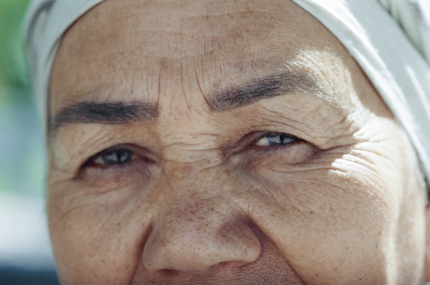 A close-up of an elderly person's face, highlighting detailed features such as wrinkles and a gentle expression. The skin tone is warm, and the person appears to be wearing a light-colored headscarf.