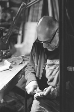 An elderly man with glasses and a bald head is focused intently on working with his hands at a task. He is wearing a dark jacket with a lighter colored patch on the chest. The man is sitting at a workbench cluttered with various tools and materials, his attention directed downwards. The background is blurry, highlighting the workshop setting.