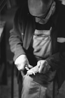 A man is engaged in a detailed craft, using tools to work on an object while seated. He is wearing glasses and an apron. The scene is captured in black and white, suggesting an artisanal or vintage setting.