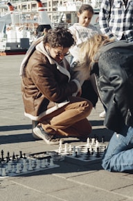 A group of people is gathered around a chess game on the pavement. One person kneels closely observing the board, wearing a brown jacket and sunglasses. A young person stands nearby, watching the game attentively. The background shows boats docked at a marina.