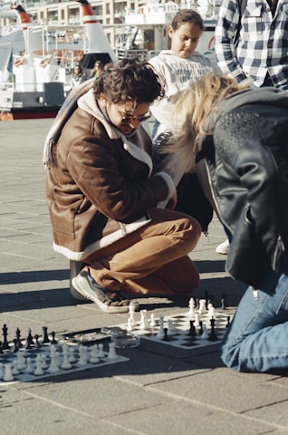 A group of people is gathered around a chess game on the pavement. One person kneels closely observing the board, wearing a brown jacket and sunglasses. A young person stands nearby, watching the game attentively. The background shows boats docked at a marina.
