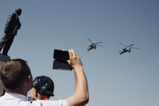 A person holds up a phone to capture two helicopters flying past a large statue. The scene suggests a public display or event, with clear blue skies creating a backdrop for the silhouettes.