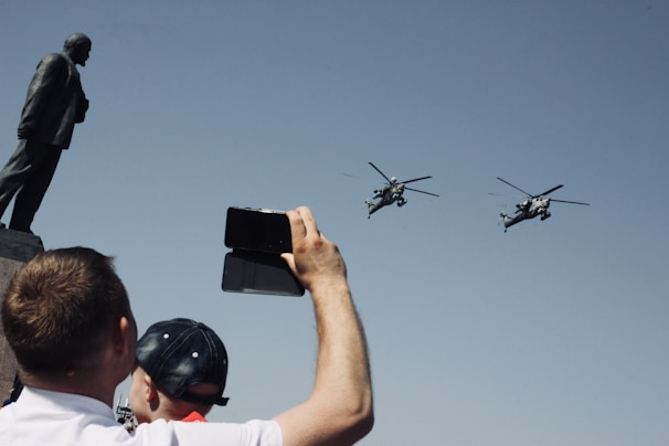 A person holds up a phone to capture two helicopters flying past a large statue. The scene suggests a public display or event, with clear blue skies creating a backdrop for the silhouettes.