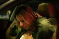 A person sitting in the driver's seat of a car, with short hair illuminated by red and green lighting. The individual is adjusting their hair, wearing a necklace and a green jacket. The overall setting is dimly lit, creating a moody atmosphere.