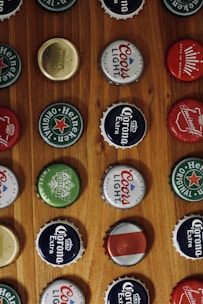 A collection of various bottle caps neatly arranged on a wooden surface, each featuring different brand logos and colors. The caps include recognizable brands such as Heineken, Coors Light, and Corona Extra, among others.