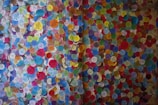 A vibrant display of colorful circular stickers densely packed on a wall, each with various symbols and text. The stickers are of multiple colors, including blue, yellow, red, green, and pink, creating a mosaic-like appearance.