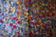 A vibrant display of colorful circular stickers densely packed on a wall, each with various symbols and text. The stickers are of multiple colors, including blue, yellow, red, green, and pink, creating a mosaic-like appearance.