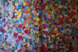 A vibrant display of colorful circular stickers densely packed on a wall, each with various symbols and text. The stickers are of multiple colors, including blue, yellow, red, green, and pink, creating a mosaic-like appearance.
