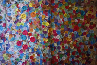 A vibrant display of colorful circular stickers densely packed on a wall, each with various symbols and text. The stickers are of multiple colors, including blue, yellow, red, green, and pink, creating a mosaic-like appearance.