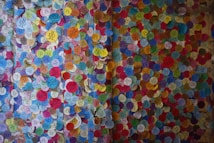 A vibrant display of colorful circular stickers densely packed on a wall, each with various symbols and text. The stickers are of multiple colors, including blue, yellow, red, green, and pink, creating a mosaic-like appearance.