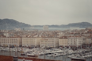 A coastal cityscape featuring a densely packed area of buildings with terra-cotta roofs. In the foreground, a busy marina is filled with numerous boats and yachts. The scene is set against a backdrop of rugged, misty mountains under an overcast sky.