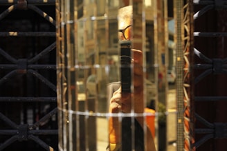 A reflection of a person holding a camera is seen in a distorted, grid-like mirrored surface. The background includes parts of an urban setting with buildings and blurred vehicles. The image incorporates various textures, including metal bars and reflective surfaces.