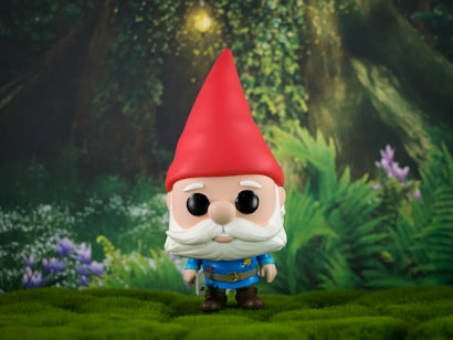 A whimsical garden gnome figure with a large red hat and blue outfit stands on a grassy surface. In the background, there is lush greenery with ferns and purple flowers under a canopy of trees, providing a serene and enchanted forest setting.