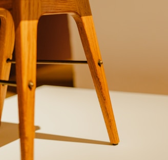 A close-up view of the legs of a wooden chair, showcasing its smooth finish and warm wood tone. The legs are angular, with visible screws and a metal support bar running between them. The surface on which the chair rests has a soft shadow cast by the legs, adding a subtle depth.