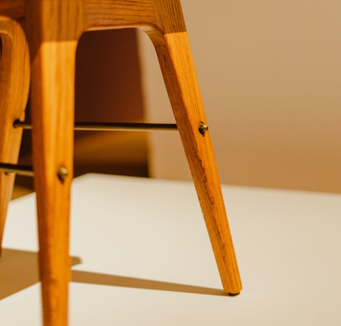 A close-up view of the legs of a wooden chair, showcasing its smooth finish and warm wood tone. The legs are angular, with visible screws and a metal support bar running between them. The surface on which the chair rests has a soft shadow cast by the legs, adding a subtle depth.