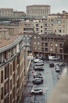 Urban scene featuring multiple residential and commercial buildings with a mix of architectural styles. A street runs through the middle with parked cars and a few people walking. The sky is overcast, casting a muted light over the scene.