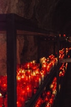 Numerous red and white candles are arranged on a series of metal shelves inside a dimly lit space, possibly a cave or a secluded grotto. The soft glow from the candles illuminates the rugged stone walls, creating a serene and contemplative atmosphere.