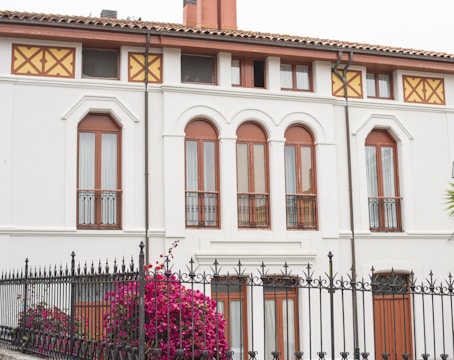 A well-maintained, two-story building displays a classic architectural style with large arched windows framed in wood and decorative wrought-iron railings. A tall wrought-iron fence encloses the front yard, which features vibrant pink bougainvillea flowers in full bloom, adding a touch of color against the white façade.