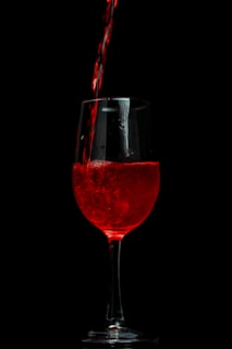 A stream of red liquid is being poured into a transparent glass against a dark background. The liquid creates a vibrant contrast with the black surroundings, highlighting the texture and color of the wine.