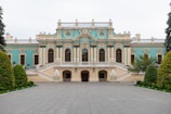 An elegant, historic palace with ornate architectural details featuring large arched windows, decorative carvings, and pastel blue and beige colors. The building is flanked by well-manicured gardens with lush greenery and symmetrical pathways leading to the entrance.