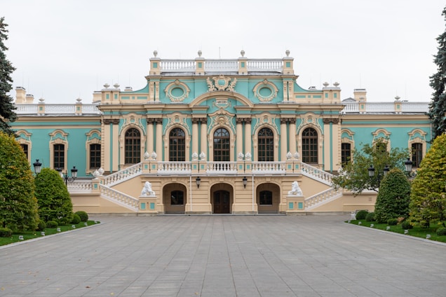 An elegant, historic palace with ornate architectural details featuring large arched windows, decorative carvings, and pastel blue and beige colors. The building is flanked by well-manicured gardens with lush greenery and symmetrical pathways leading to the entrance.