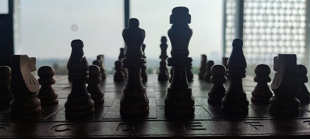 A silhouette of wooden chess pieces arranged on a chessboard, set against a bright window, creating a stark contrast between light and dark.