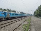 A long train with blue carriages travels on a set of parallel railway tracks. Overhead power lines are visible, suggesting an electrified rail system. The scene is set in an outdoor environment with a line of trees and greenery alongside the track. The sky is overcast, giving the setting a muted tone. A platform on the right appears empty, with a few benches placed along it.