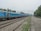 A long train with blue carriages travels on a set of parallel railway tracks. Overhead power lines are visible, suggesting an electrified rail system. The scene is set in an outdoor environment with a line of trees and greenery alongside the track. The sky is overcast, giving the setting a muted tone. A platform on the right appears empty, with a few benches placed along it.