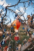 A tree is adorned with numerous blue evil eye charms and a colorful hot air balloon ornament, creating a visually intricate display. The charms are predominantly round with a white and light blue center, contrasted by the vibrant red, yellow, and orange hues of the balloon. The background is set against a blue sky with some clouds, adding a natural and serene element.