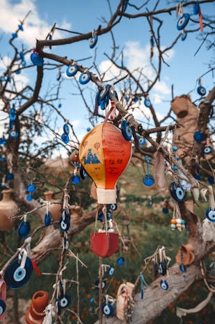 A tree is adorned with numerous blue evil eye charms and a colorful hot air balloon ornament, creating a visually intricate display. The charms are predominantly round with a white and light blue center, contrasted by the vibrant red, yellow, and orange hues of the balloon. The background is set against a blue sky with some clouds, adding a natural and serene element.