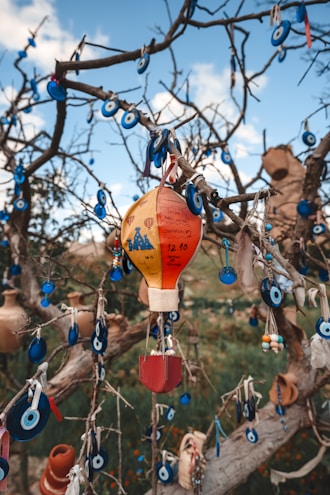 A tree is adorned with numerous blue evil eye charms and a colorful hot air balloon ornament, creating a visually intricate display. The charms are predominantly round with a white and light blue center, contrasted by the vibrant red, yellow, and orange hues of the balloon. The background is set against a blue sky with some clouds, adding a natural and serene element.