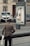 An elderly person with a cane approaches a kiosk on a sidewalk, where the display panel is significantly damaged. The background shows parked cars and a residential building with a door and windows.