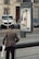 An elderly person with a cane approaches a kiosk on a sidewalk, where the display panel is significantly damaged. The background shows parked cars and a residential building with a door and windows.