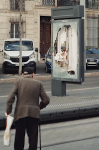 An elderly person with a cane approaches a kiosk on a sidewalk, where the display panel is significantly damaged. The background shows parked cars and a residential building with a door and windows.