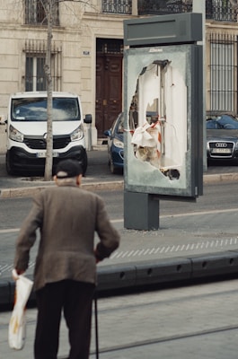 An elderly person with a cane approaches a kiosk on a sidewalk, where the display panel is significantly damaged. The background shows parked cars and a residential building with a door and windows.