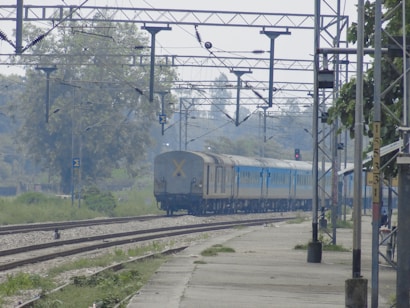 A blue passenger train is moving along the railway tracks under an overhead electric line infrastructure. The train is passing through a rural area surrounded by trees and greenery. Electrical poles and cables are visible above the train, and part of a railway platform is seen on the right side.