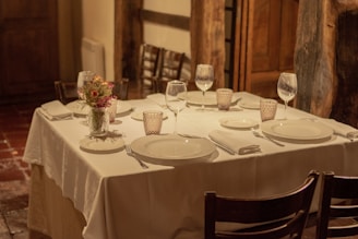 A neatly set dining table with a white tablecloth featuring plates, wine glasses, cutlery, and cloth napkins. A small bouquet of flowers serves as the centerpiece. The setting appears to be cozy, with wooden elements and a warm, dimly-lit ambiance.