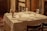 A neatly set dining table with a white tablecloth featuring plates, wine glasses, cutlery, and cloth napkins. A small bouquet of flowers serves as the centerpiece. The setting appears to be cozy, with wooden elements and a warm, dimly-lit ambiance.