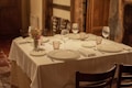 A neatly set dining table with a white tablecloth featuring plates, wine glasses, cutlery, and cloth napkins. A small bouquet of flowers serves as the centerpiece. The setting appears to be cozy, with wooden elements and a warm, dimly-lit ambiance.