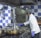 A chef wearing a white coat and purple gloves is cooking in a commercial kitchen. The kitchen features a blue and white checkered wall, multiple metal pots on a stove, and an industrial oven. The chef appears focused on stirring the contents of one of the pots.