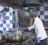 A chef wearing a white coat and purple gloves is cooking in a commercial kitchen. The kitchen features a blue and white checkered wall, multiple metal pots on a stove, and an industrial oven. The chef appears focused on stirring the contents of one of the pots.