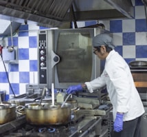 A chef wearing a white coat and purple gloves is cooking in a commercial kitchen. The kitchen features a blue and white checkered wall, multiple metal pots on a stove, and an industrial oven. The chef appears focused on stirring the contents of one of the pots.