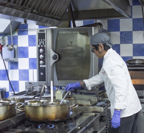 A chef wearing a white coat and purple gloves is cooking in a commercial kitchen. The kitchen features a blue and white checkered wall, multiple metal pots on a stove, and an industrial oven. The chef appears focused on stirring the contents of one of the pots.