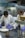 Two chefs in a professional kitchen are focused on preparing dishes. One chef is plating a dish with tomato sauce while the other is spooning sauce over a piece of meat. They are wearing white chef coats and blue gloves.