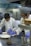 Two chefs in a professional kitchen are focused on preparing dishes. One chef is plating a dish with tomato sauce while the other is spooning sauce over a piece of meat. They are wearing white chef coats and blue gloves.