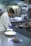 A chef in a white coat is preparing food in a commercial kitchen. He is wearing blue gloves and plating a dish on a stainless steel counter. The kitchen is equipped with industrial appliances including a stove and refrigerators.