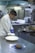 A chef in a white coat is preparing food in a commercial kitchen. He is wearing blue gloves and plating a dish on a stainless steel counter. The kitchen is equipped with industrial appliances including a stove and refrigerators.