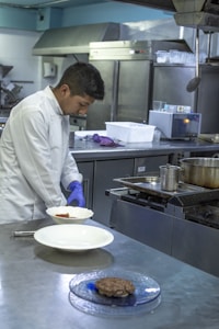 A chef in a white coat is preparing food in a commercial kitchen. He is wearing blue gloves and plating a dish on a stainless steel counter. The kitchen is equipped with industrial appliances including a stove and refrigerators.